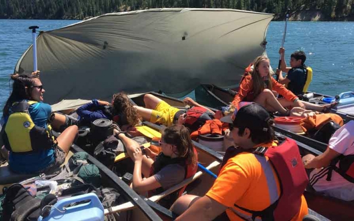 a group of canoes tethered together hold high school students as they use their paddles to prop up a tarp for shade